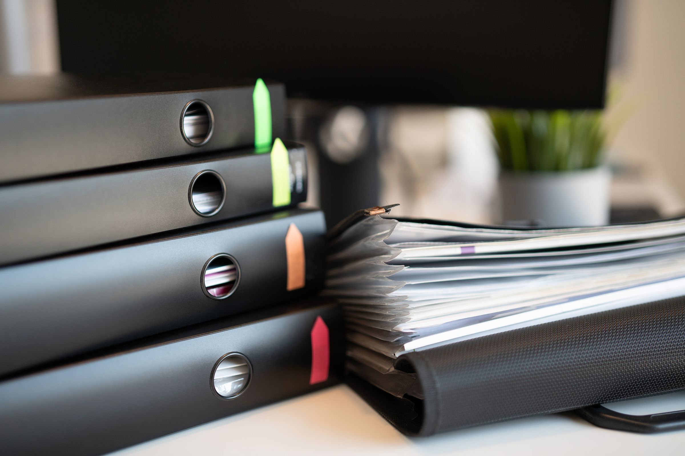 A stack of black ring binders marked with coloured index tabs beside a thick file of documents on an office desk