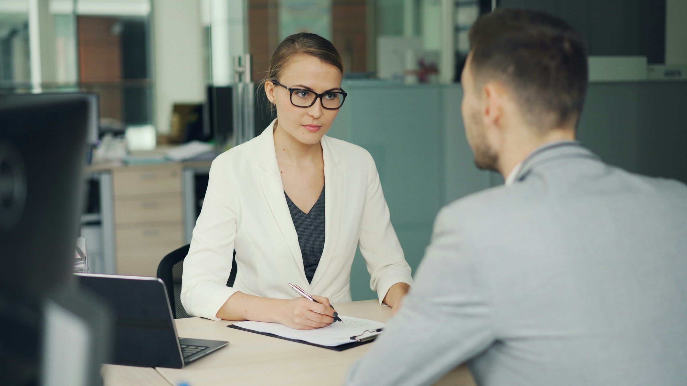 An adviser in glasses and a white blazer listens and takes notes on a clipboard while a client in a grey jacket, seen from behind, talks with her across an office desk
