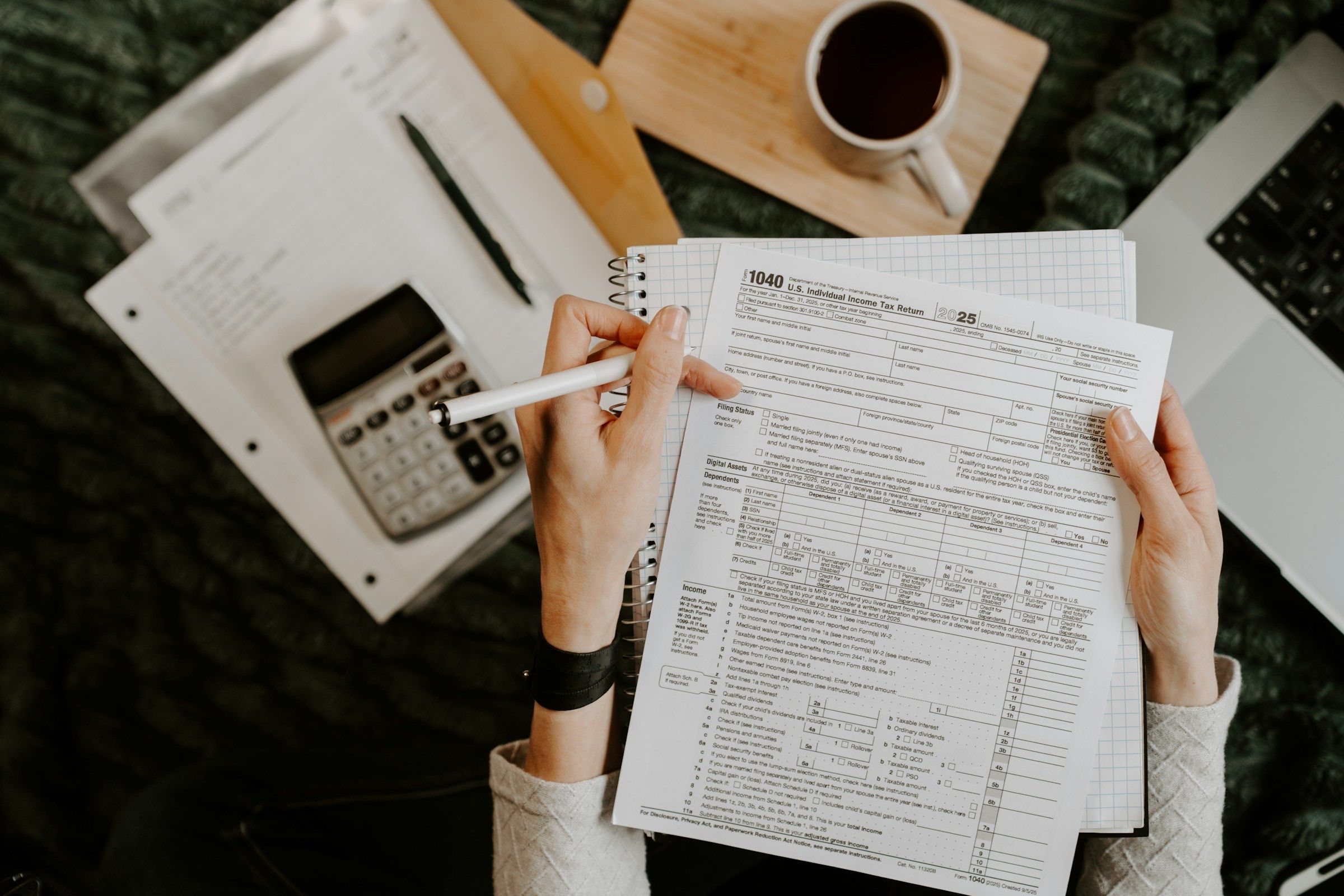 Overhead view of hands holding a blank 2025 US Form 1040 tax return and a white pen, beside a calculator, a cup of black coffee and a laptop on a dark green knit blanket