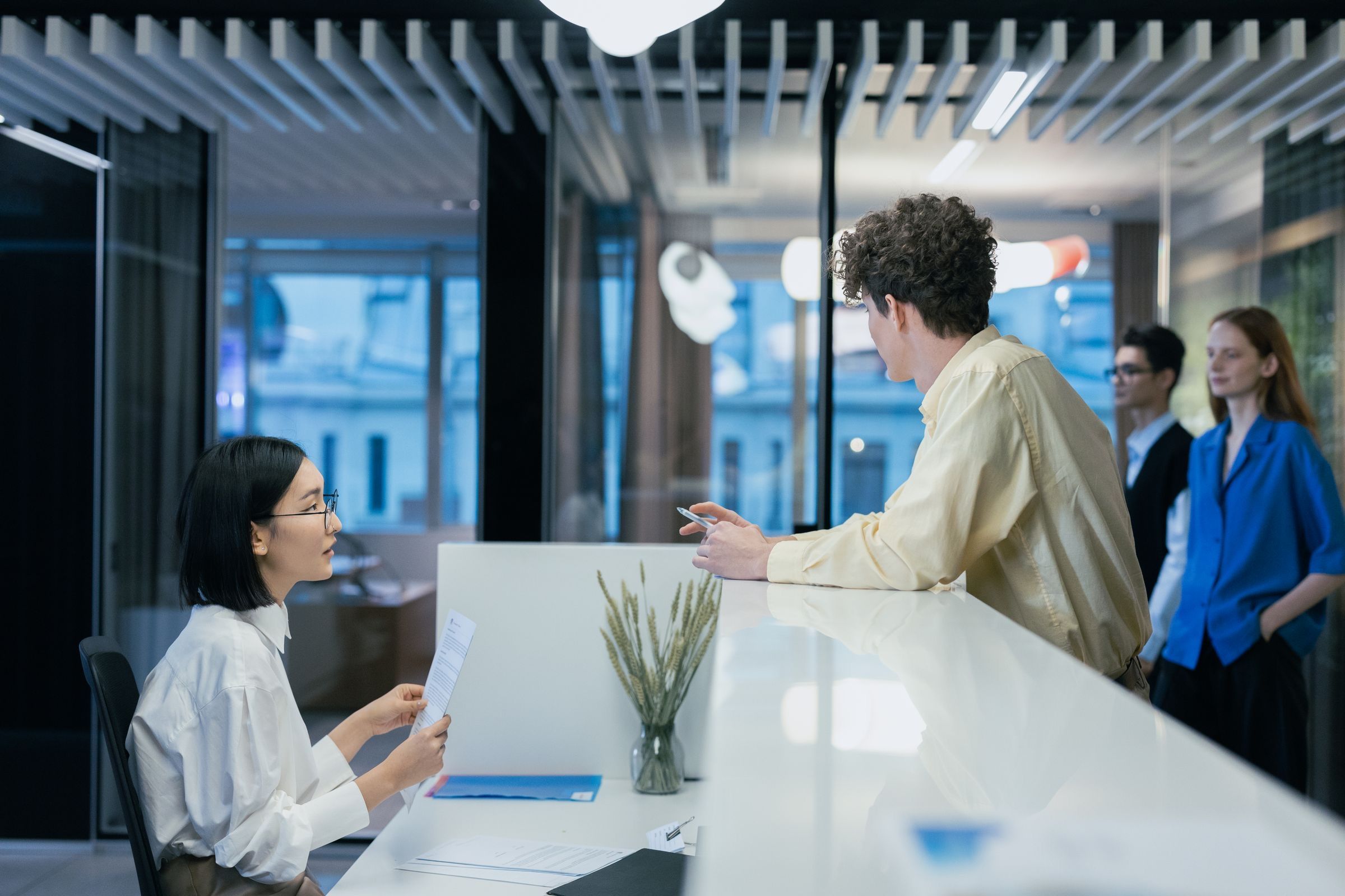 A receptionist in a white shirt holds a letter as she talks with a visitor leaning on a white front desk in a glass-walled office