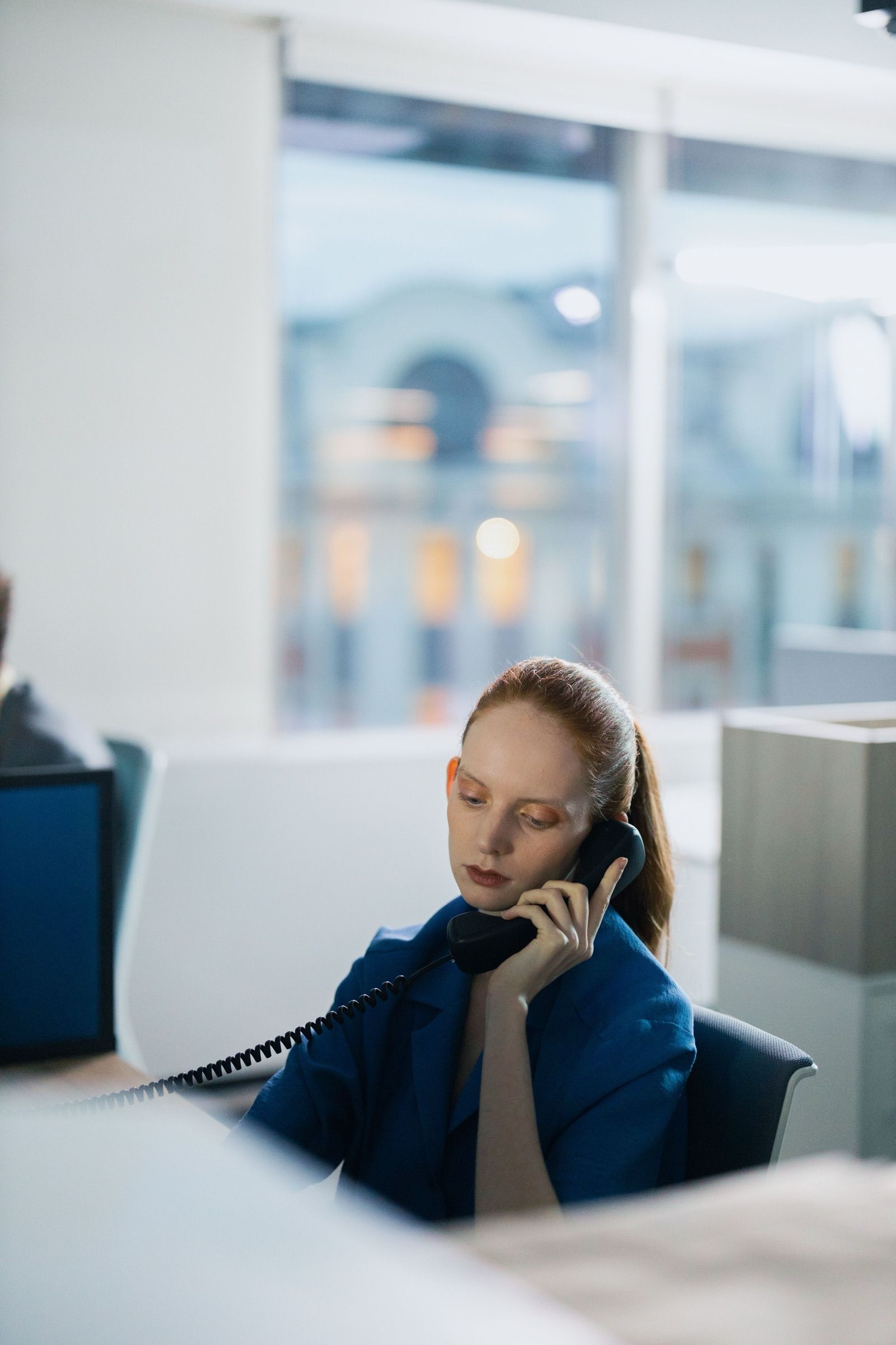 A woman in a cobalt-blue jacket listens on a corded desk phone at her workstation in a bright, glass-walled office with a blurred building behind the window